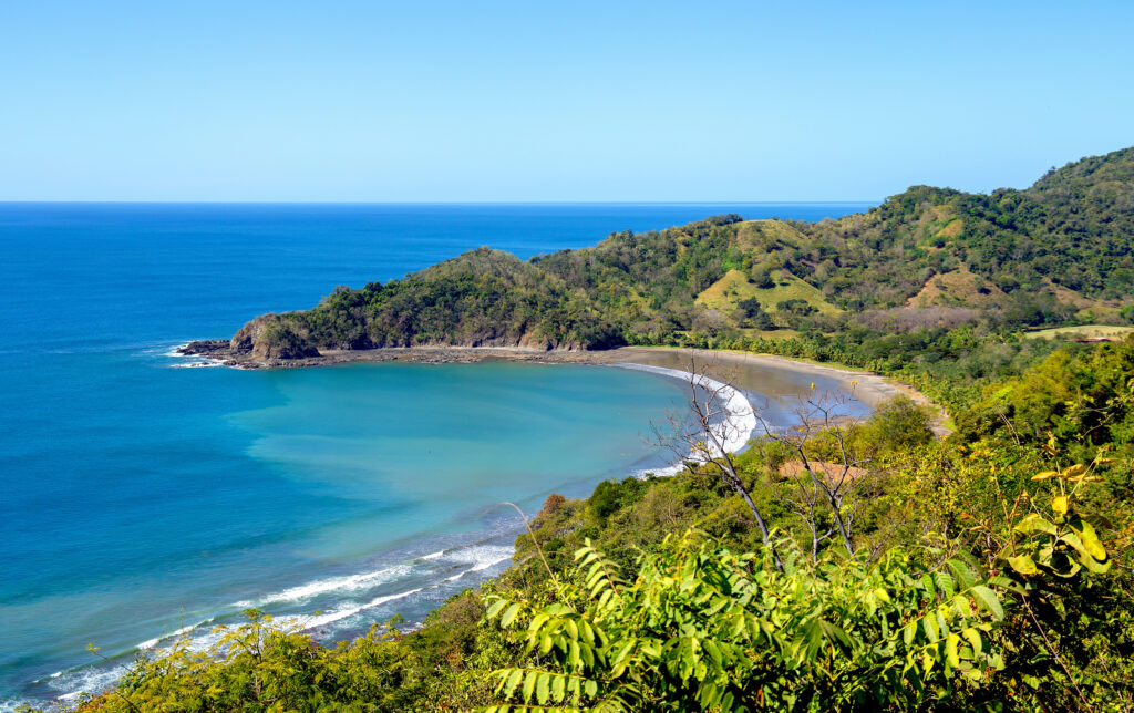 Bird's eye view of the landscape of Punta Islita with turquoise sea, dark sandy beach and lush, green hilly landscape. Bushy plants in the foreground, a tropical lonesome bay in Costa Rica