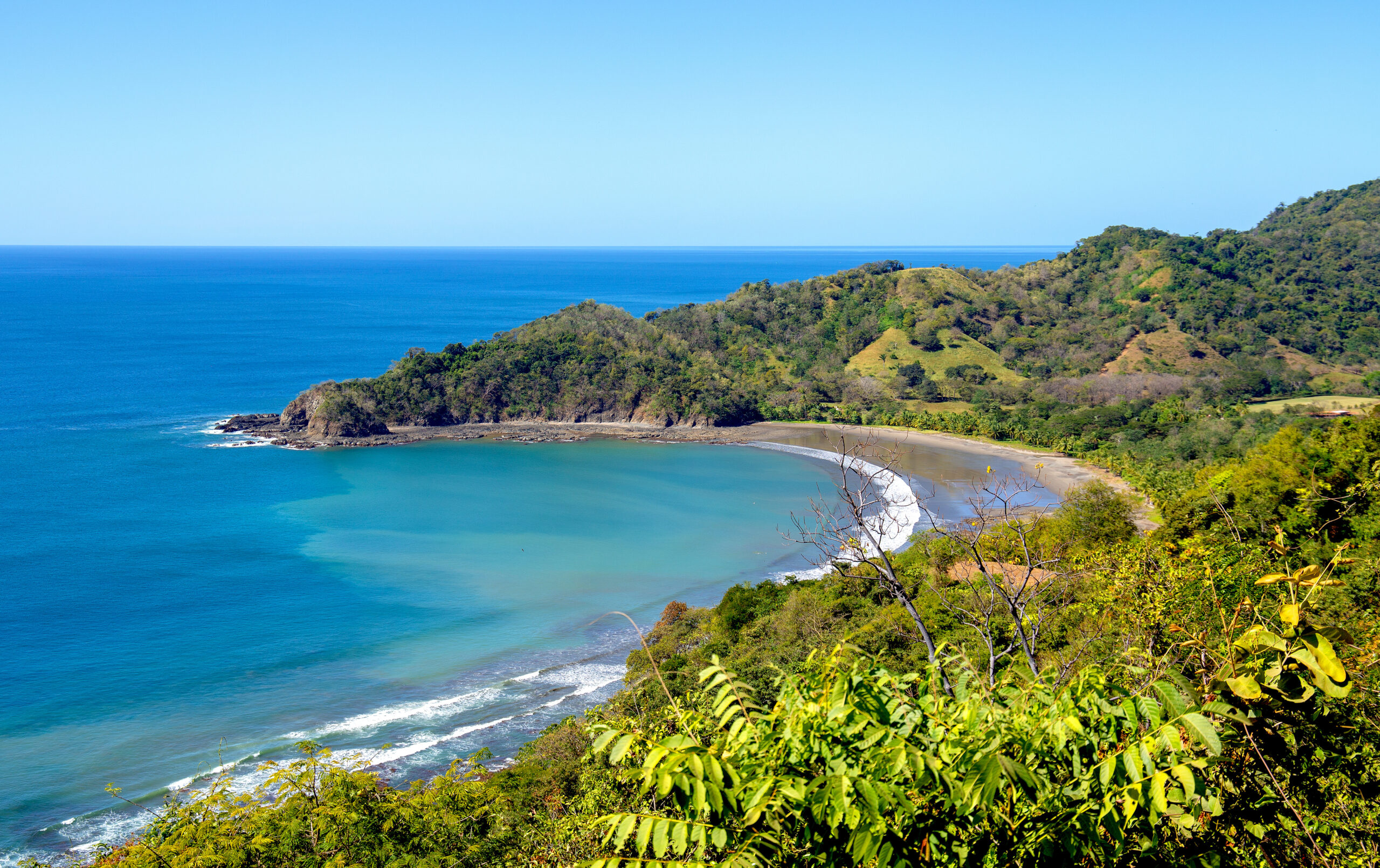 Bird's eye view of the landscape of Punta Islita with turquoise sea, dark sandy beach and lush, green hilly landscape. Bushy plants in the foreground, a tropical lonesome bay in Costa Rica